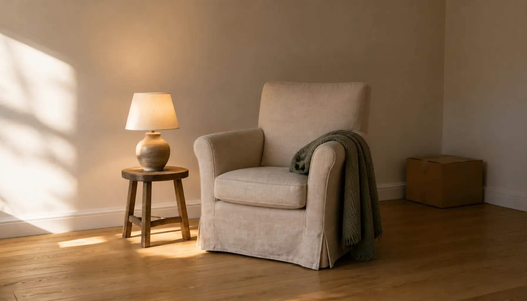 A warm table lamp beside a linen armchair in a bare cream room with a sage throw and soft afternoon light from the left